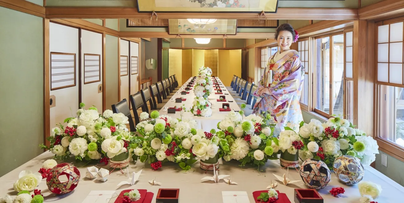 テーブルの上に花束が飾られ、奥に金屏風がある和室の会場に着物姿の花嫁が立っている